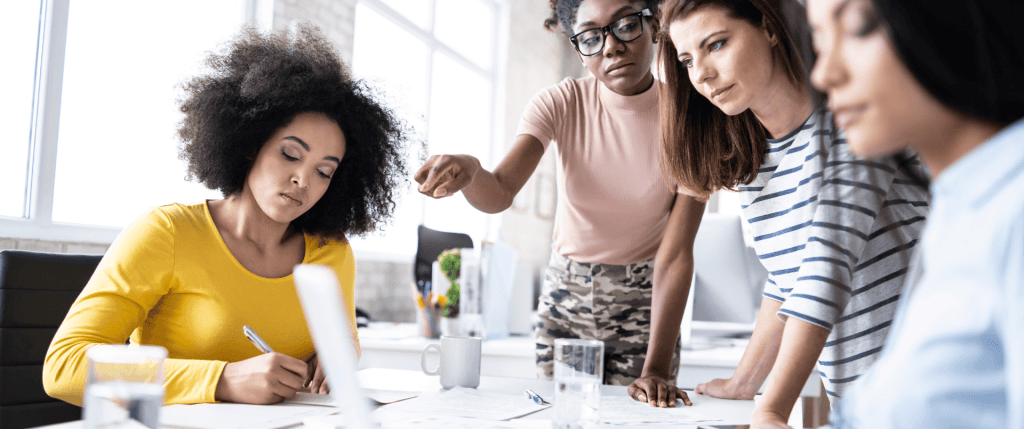 A diverse group of four women collaborating at a table, with one woman writing notes while the others observe and discuss.